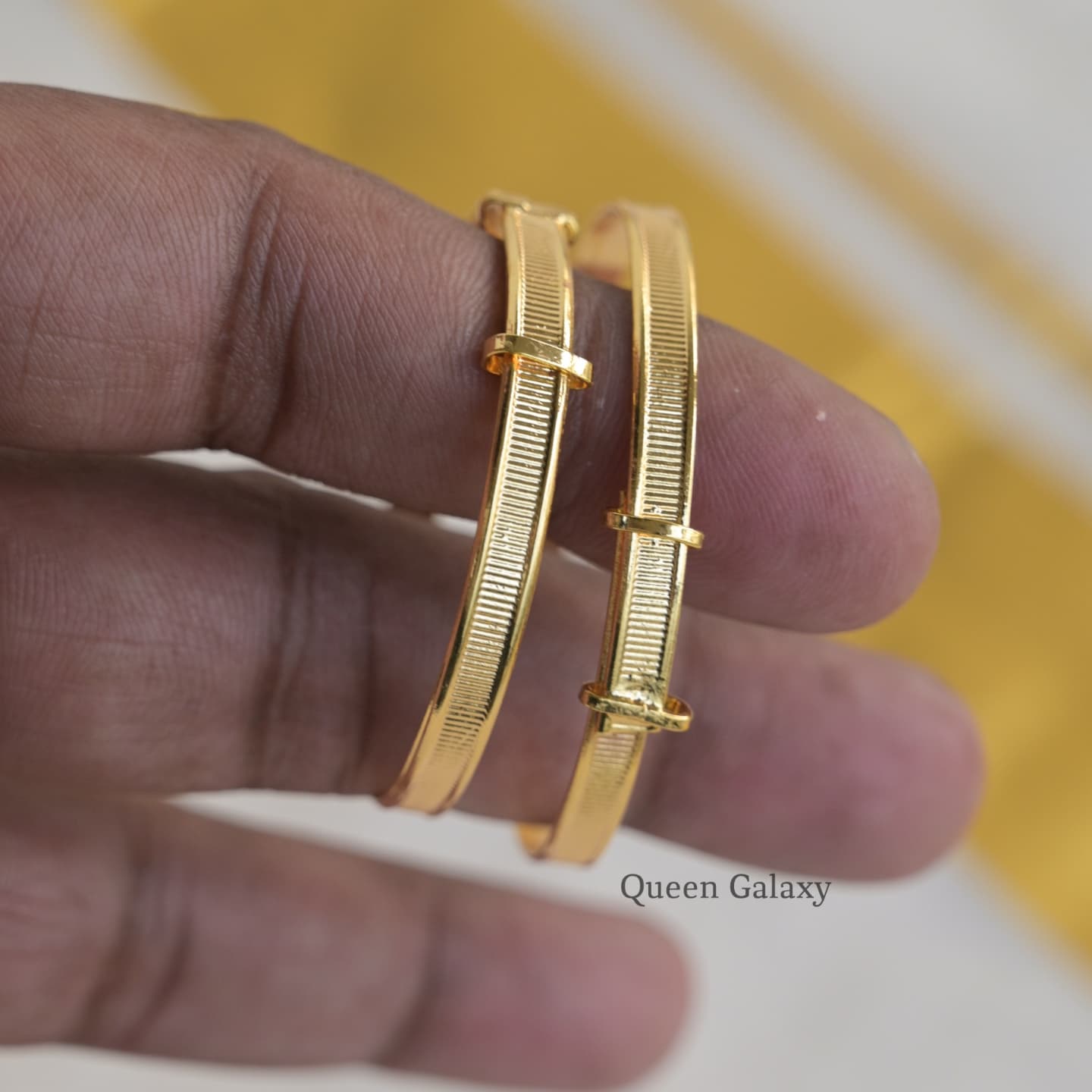 Two gold-plated baby bangles held between fingers, with a yellow background.