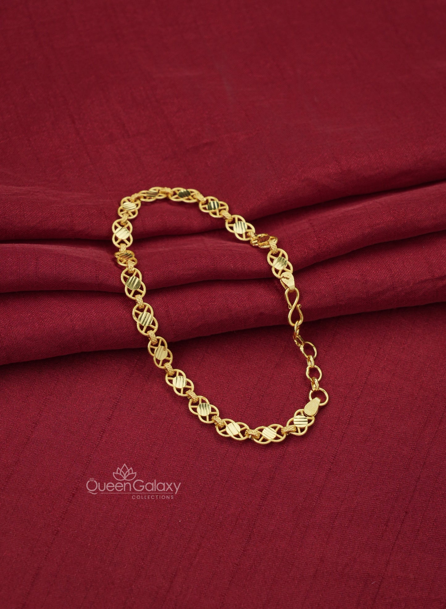 A gold plated crisscross chain bracelet with a patterned design, displayed on a red fabric surface at queen galaxy collections