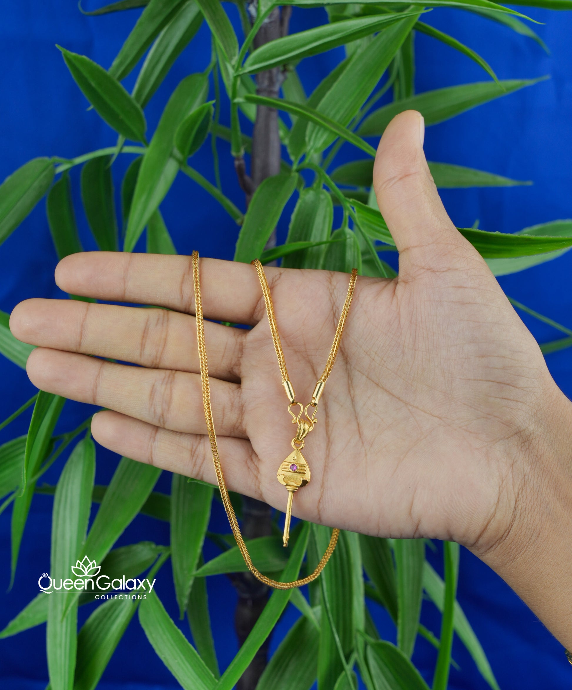 Gold plated box chain with murukan vel pendant held in a hand with a blue background and plant