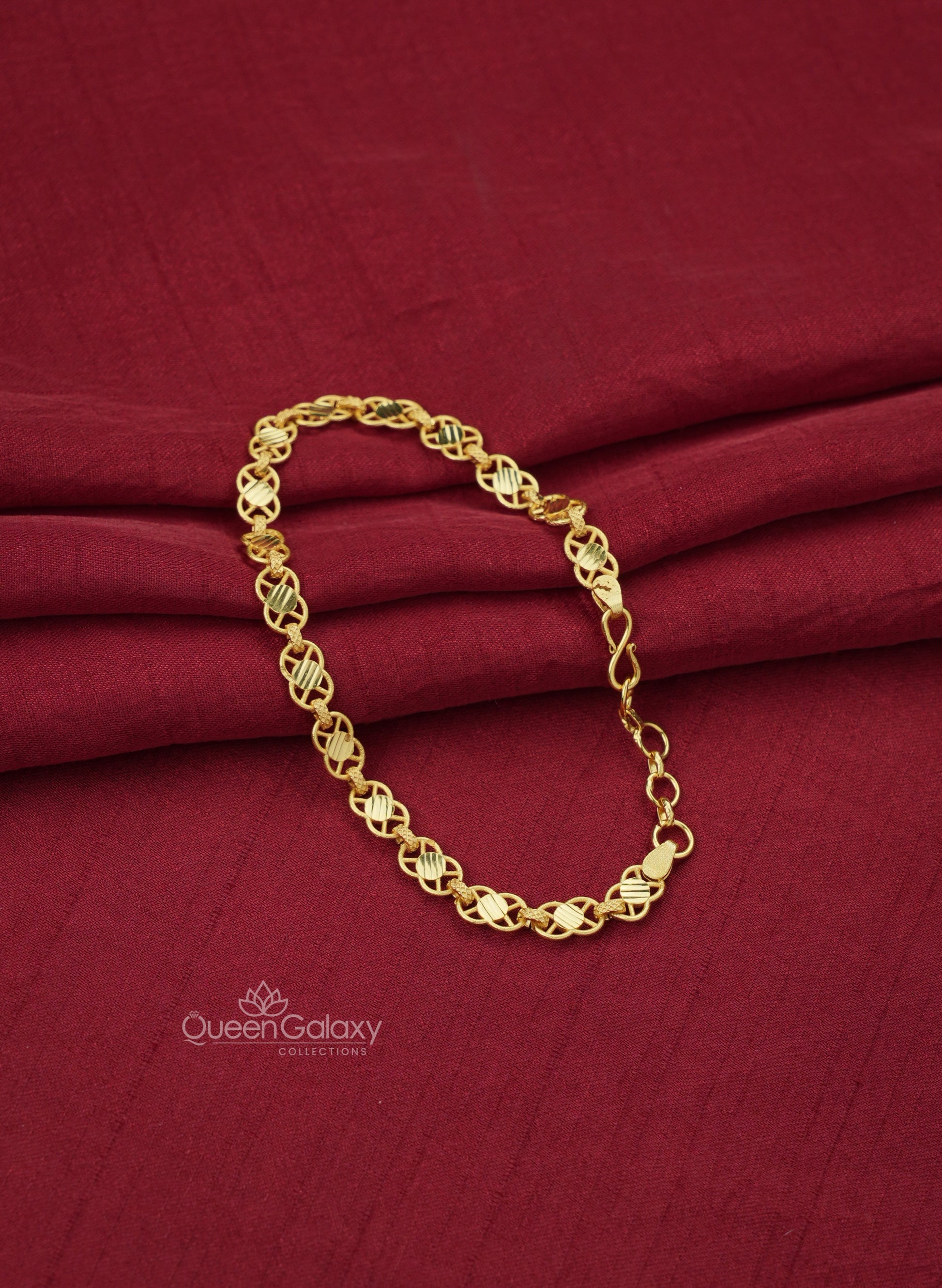 A gold plated crisscross chain bracelet with a patterned design, displayed on a red fabric surface at queen galaxy collections